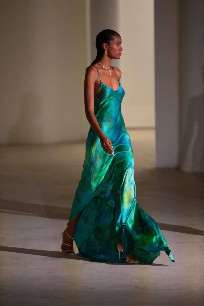 A model wearing a blue-green flowing slip dress on the runway.