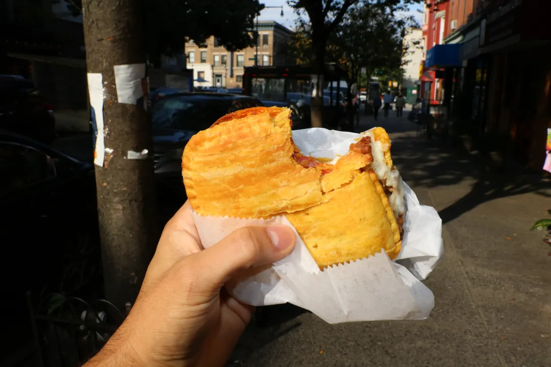 Hand holding a Jamaican beef patty wrapped in a paper napkin, emphasizing street food ritual and immediacy