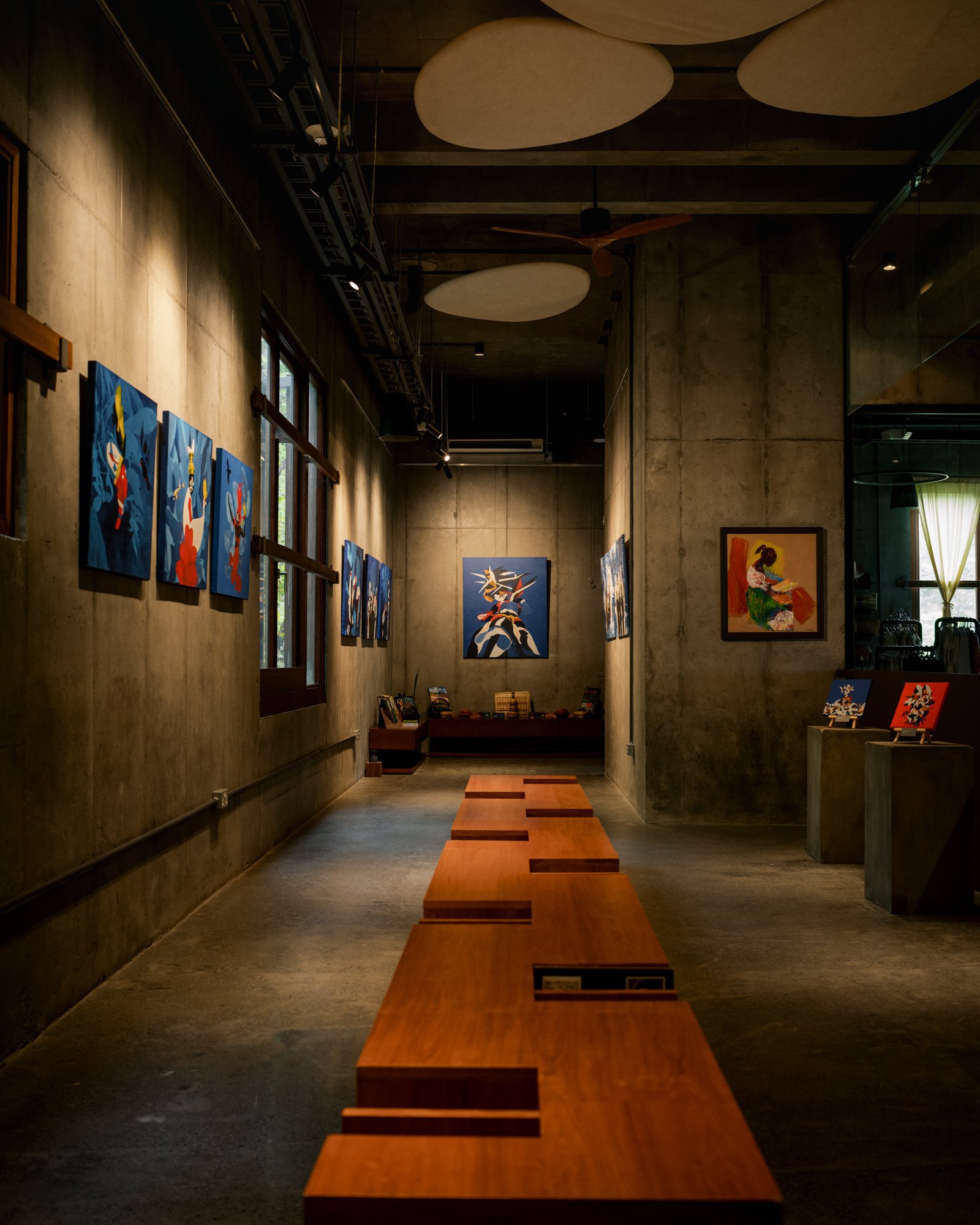 Interior gallery space with paintings on concrete walls and wooden benches.