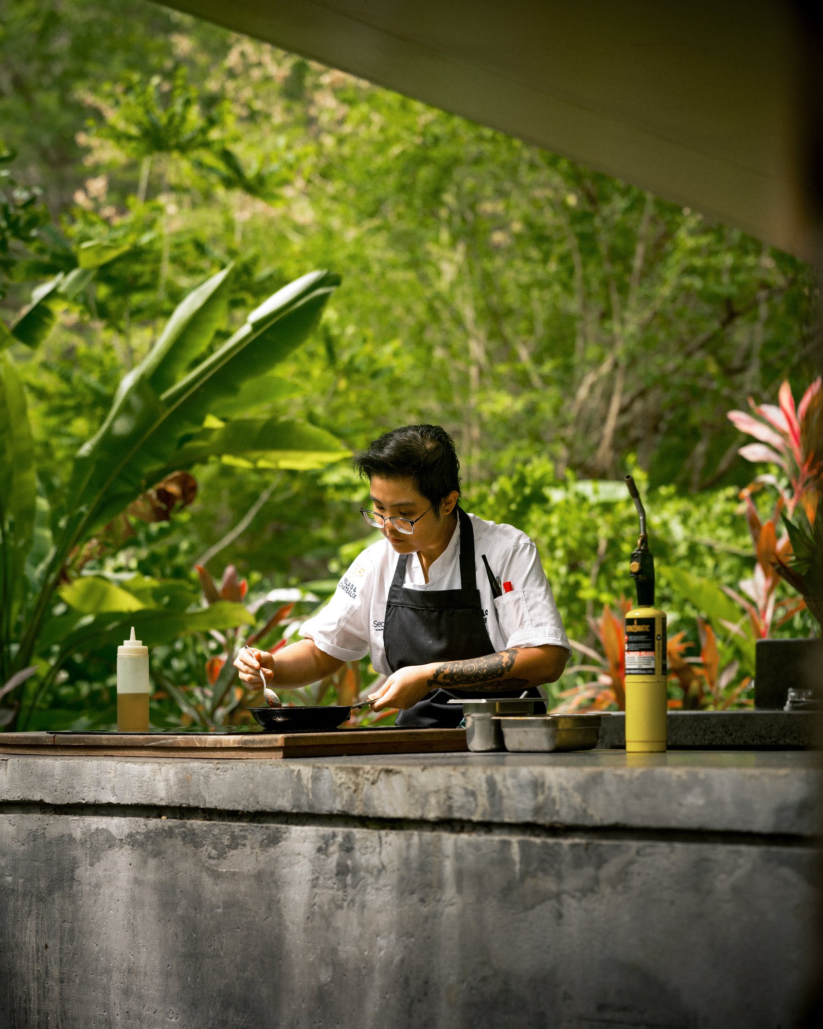 Chef preparing food at an outdoor counter surrounded by tropical greenery.
