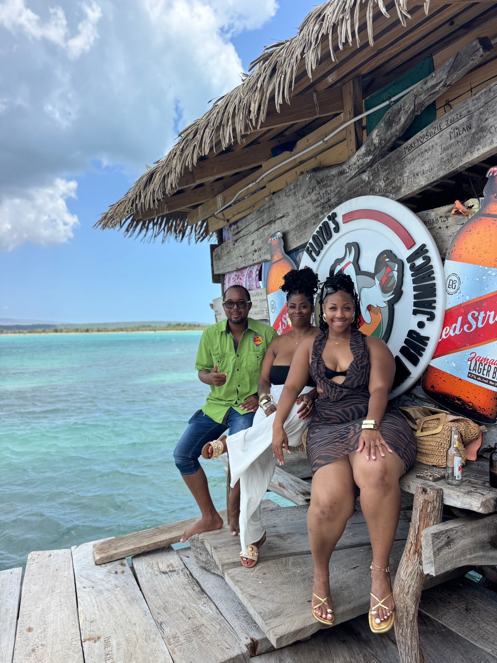 Three adults beside an overwater wooden bar with a Floyd’s Pelican Bar sign.